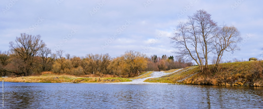 Obraz premium Landscape. Trees, dry grass and a country road along the banks of a small river in late autumn.