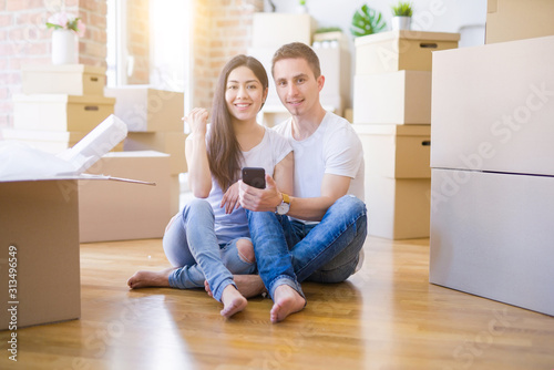 Beautiful couple sitting on the floor using smartphone at new home around cardboard boxes pointing and showing with thumb up to the side with happy face smiling © Krakenimages.com