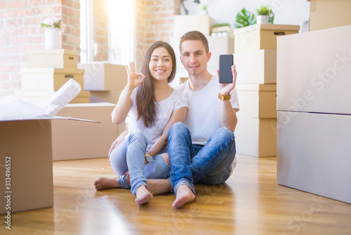 Beautiful couple sitting on the floor holding smartphone at new home around cardboard boxes doing ok sign with fingers, excellent symbol © Krakenimages.com