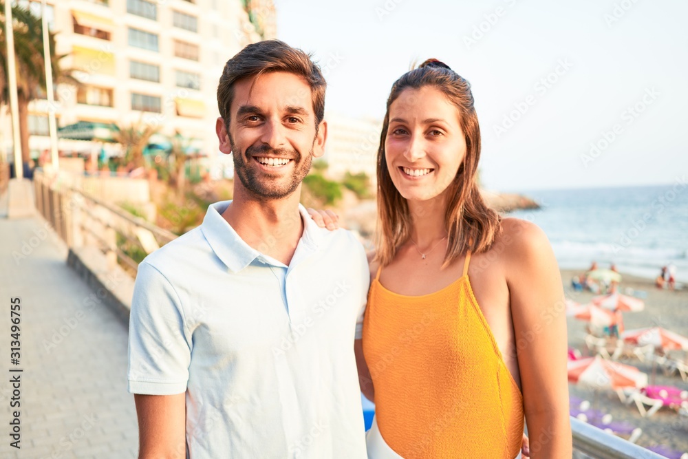 Young beautiful couple on vacation smiling happy and confident. Standing with smile on face hugging at the beach Young beautiful couple on vacation smiling happy and confident. Standing with smile on face hugging at the beach