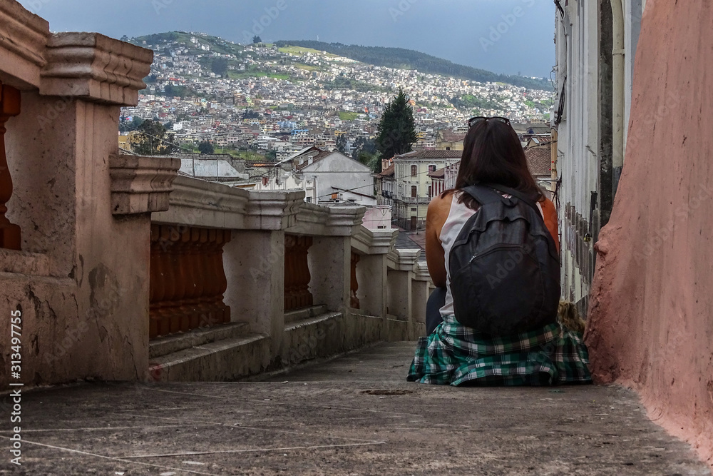 A woman sitting on old stairs of the old town of Quito, Ecuador. She is with her back turned and a beautiful mount landscape of the village can be seen behind.