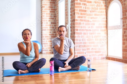 Middle age sporty couple sitting on mat doing stretching yoga exercise at gym looking at the camera blowing a kiss with hand on air being lovely and sexy. Love expression.