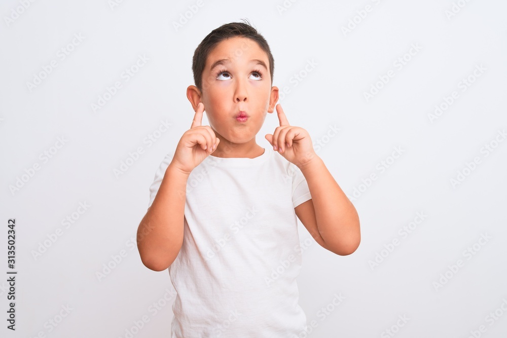 Beautiful kid boy wearing casual t-shirt standing over isolated white background amazed and surprised looking up and pointing with fingers and raised arms. Beautiful kid boy wearing casual t-shirt standing over isolated white background amazed and surprised looking up and pointing with fingers and raised arms.