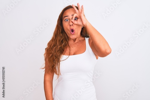 Middle age mature woman standing over white isolated background doing ok gesture shocked with surprised face, eye looking through fingers. Unbelieving expression.