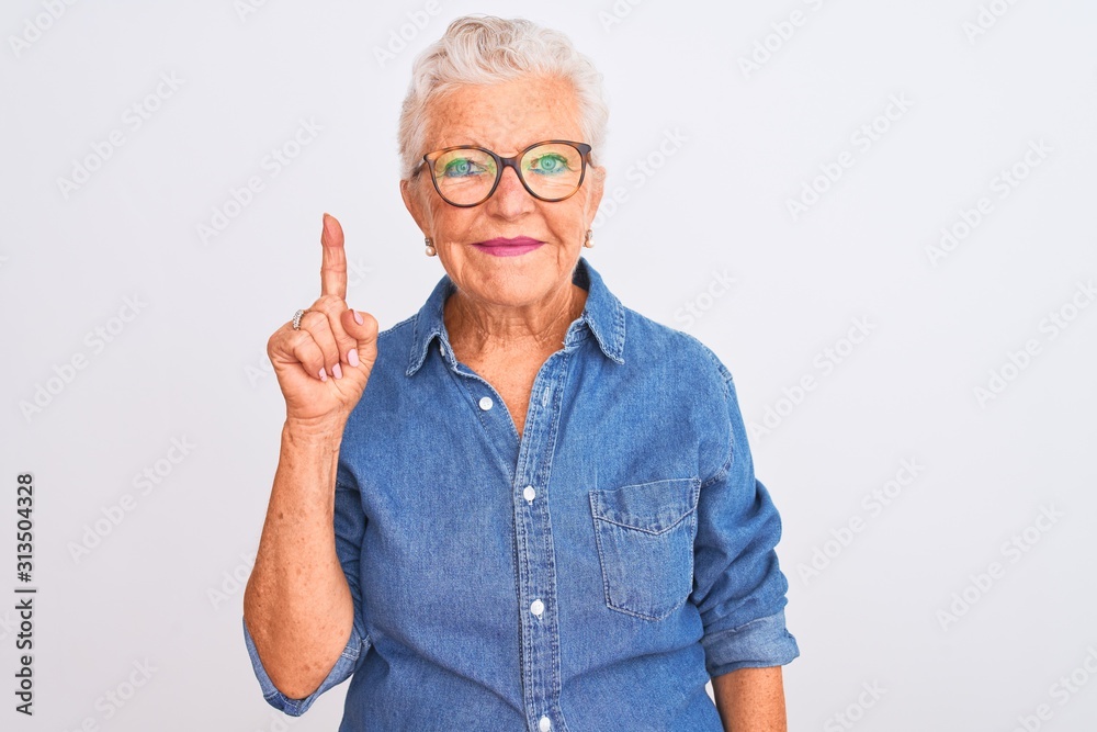 Senior grey-haired woman wearing denim shirt and glasses over isolated white background showing and pointing up with finger number one while smiling confident and happy. - obrazy, fototapety, plakaty 