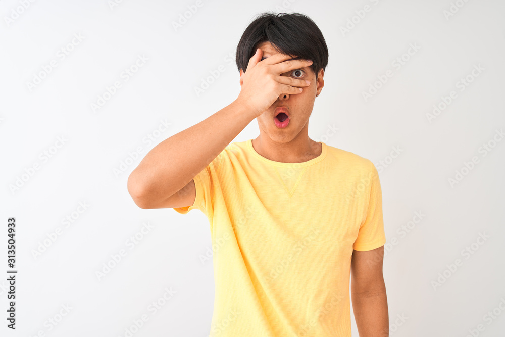 Chinese man wearing yellow casual t-shirt standing over isolated white background peeking in shock covering face and eyes with hand, looking through fingers with embarrassed expression. Chinese man wearing yellow casual t-shirt standing over isolated white background peeking in shock covering face and eyes with hand, looking through fingers with embarrassed expression.