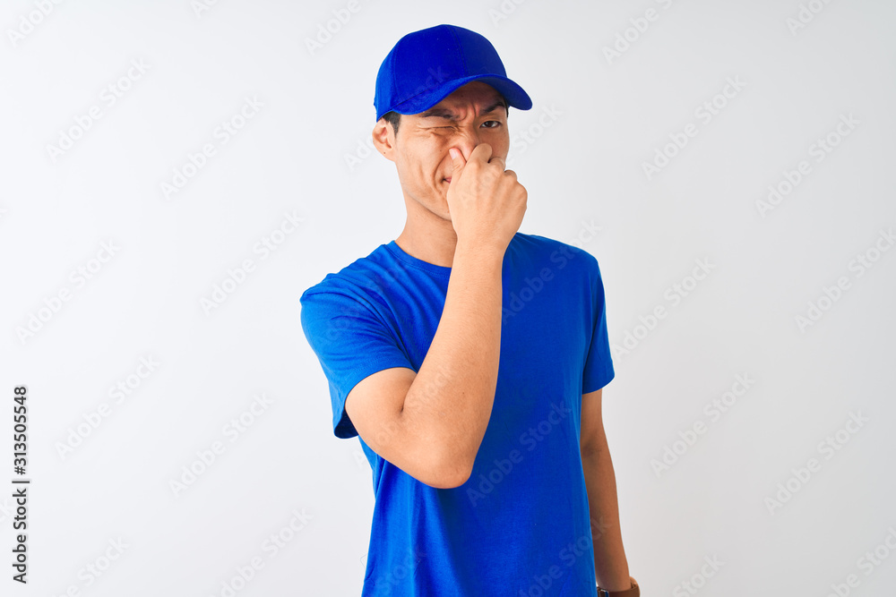 Chinese deliveryman wearing blue t-shirt and cap standing over isolated white background smelling something stinky and disgusting, intolerable smell, holding breath with fingers on nose. Bad smell