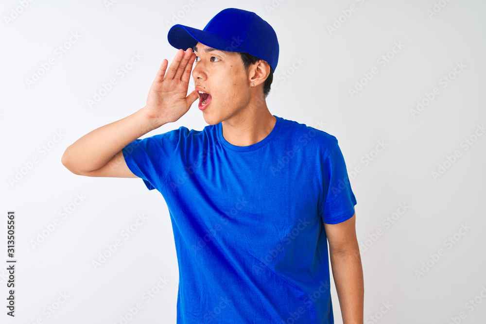 Chinese deliveryman wearing blue t-shirt and cap standing over isolated white background shouting and screaming loud to side with hand on mouth. Communication concept.