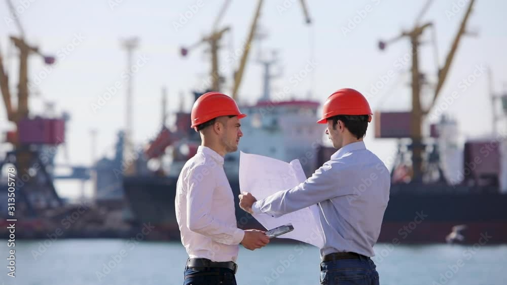 Two architects communicate and show whatman paper project on the background of a shipyard with cargo port cranes Two architects communicate and show whatman paper project on the background of a shipyard with cargo port cranes