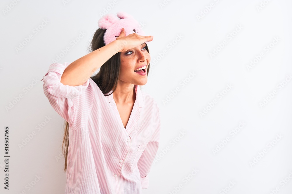 Young beautiful woman wearing sleep mask and pajama over isolated white background very happy and smiling looking far away with hand over head. Searching concept. Young beautiful woman wearing sleep mask and pajama over isolated white background very happy and smiling looking far away with hand over head. Searching concept.