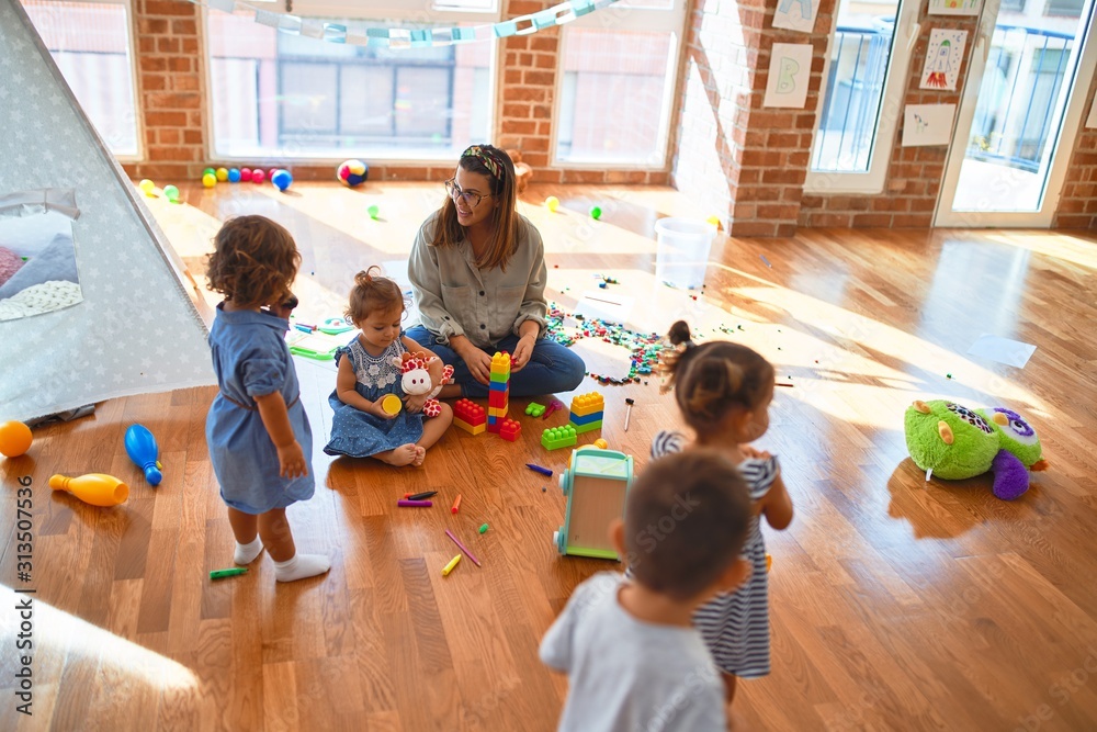 Beautiful teacher and group of toddlers playing around lots of toys at kindergarten - obrazy, fototapety, plakaty 
