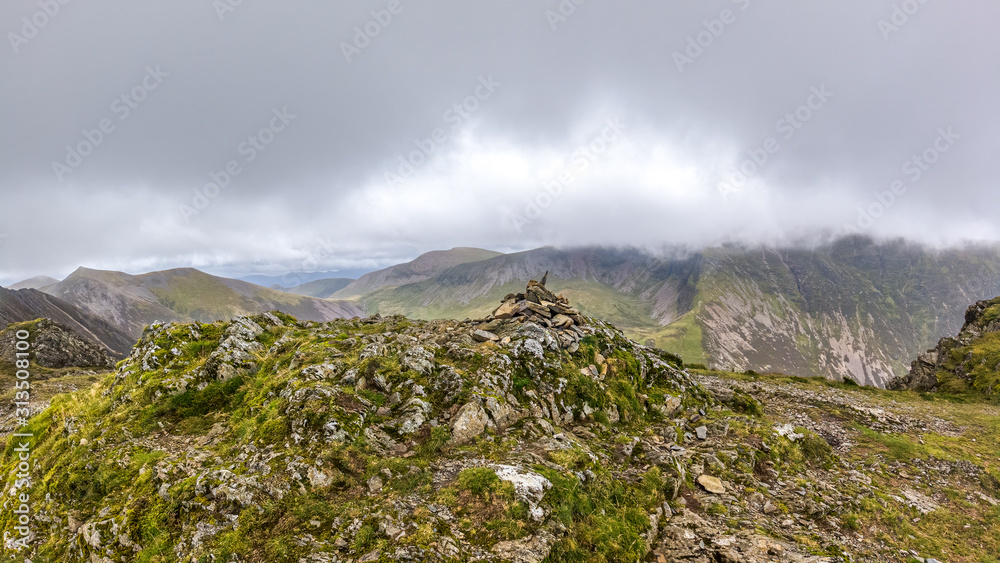 A scenic view of a rocky mountain summit with a stony cairn and mountain range in the background under a stormy grey cloudy sky
