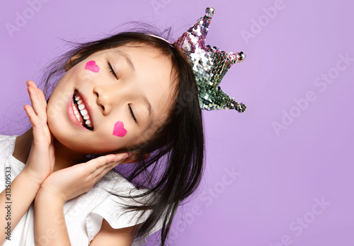 Asian Korean kid girl princess in crown closeup portrait Happy smiling laughing with a painted red hearts sign on cheeks