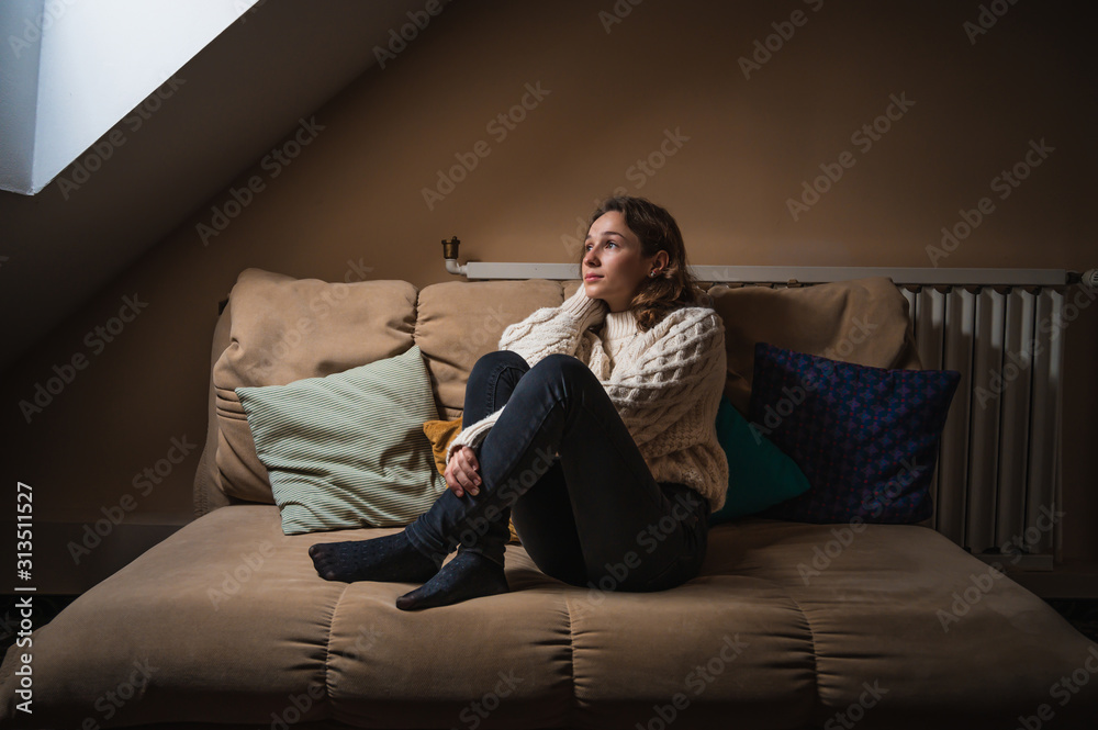 Beautiful young woman looking out the window of a room with winter clothes, resting and relaxing at home on a winter morning Beautiful young woman looking out the window of a room with winter clothes, resting and relaxing at home on a winter morning