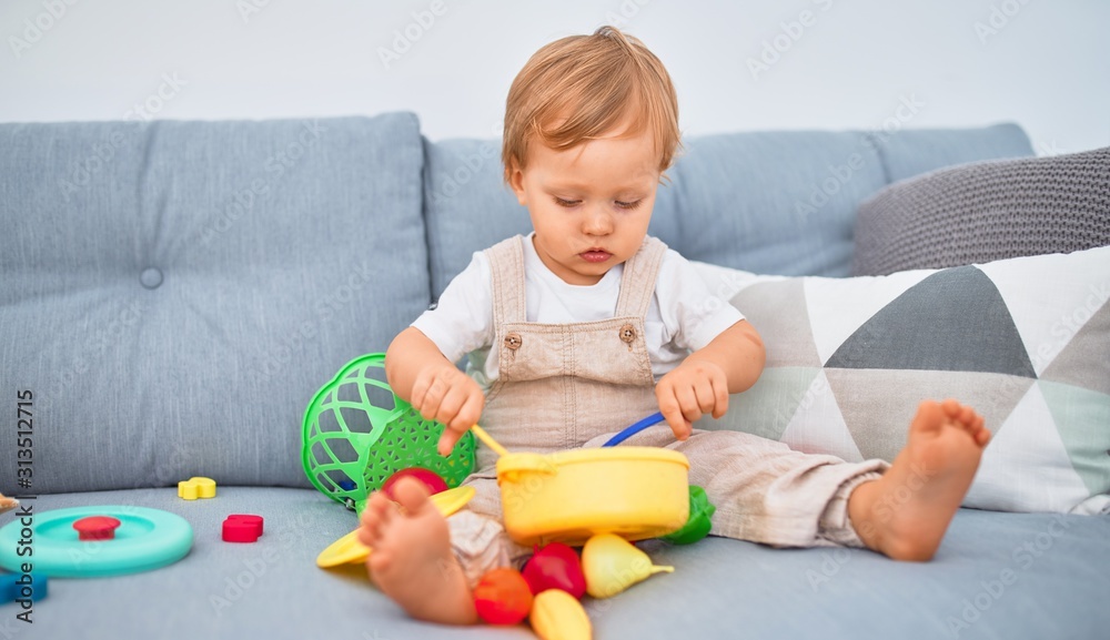 Adorable blonde toddler sitting on the sofa playing with plastic meals toys at home Adorable blonde toddler sitting on the sofa playing with plastic meals toys at home