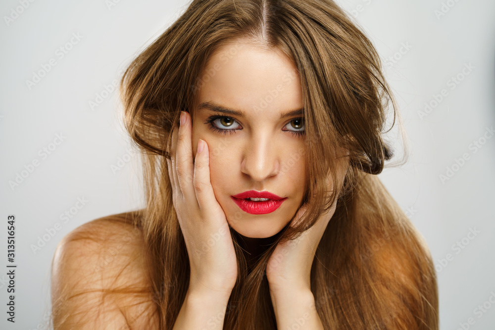 Fototapeta premium Beauty portrait of beautiful caucasian girl young brunette woman with red lipstick natural perfect skin and makeup holding hands on her chin looking to the camera in studio
