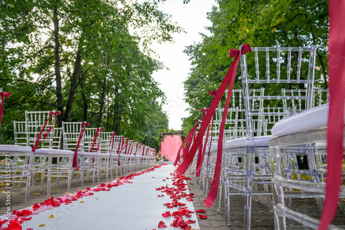 Wedding ceremony on a summer day in the city park among the green foliage. White carpet, chairs decorated with red ribbons.