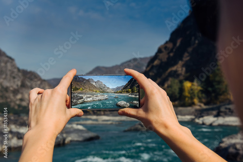 Hands hold a smartphone and take photo of mountain river and a rocks, close-up. Photographing the beautiful landscape on smartphone while traveling.