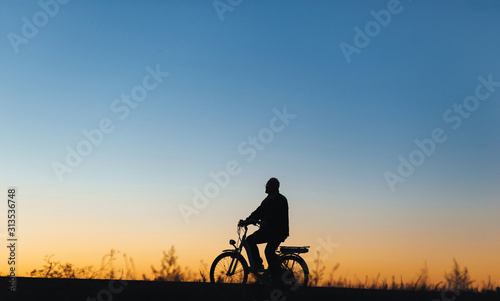 Male cyclist on the e-bike or electric bicycle on the sunset background riding up the hill. Silhouette of the old man in profile. Active pension. Travel. Sport. Copy space.