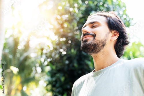 A bearded man is meditating outdoor in the park with face raised up to sky and eyes closed on sunny summer day. Concept of meditation, dreaming, wellbeing healthy lifestyle photo