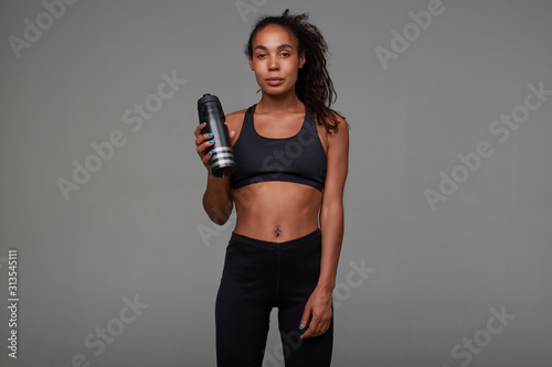 Tired pretty young dark skinned curly brunette female with casual hairstyle relaxing after hard training, posing over grey background with black fitness bottle in her arm © timtimphoto