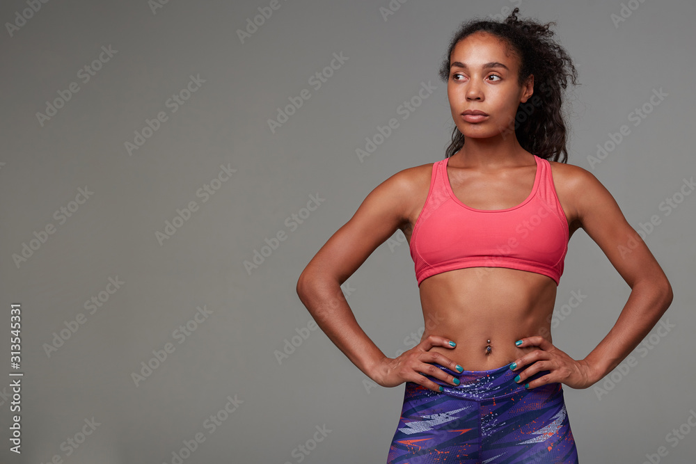 Studio shot of young attractive dark skinned curly woman with ponytail hairstyle looking aside with folded lips and keeping her hands on waist, isolated over grey background Studio shot of young attractive dark skinned curly woman with ponytail hairstyle looking aside with folded lips and keeping her hands on waist, isolated over grey background