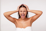 Portrait of stressed young brunette female wrapped in bath towel clutching her head with raised hands and screaming with wide mouth opened, keeping eyes closed while posing over white background