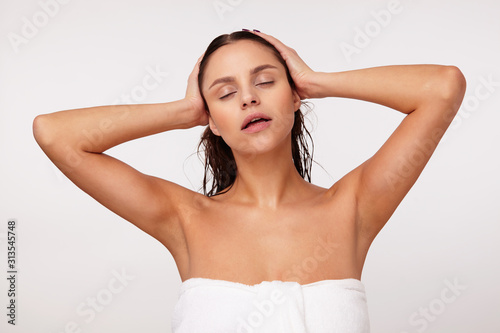 Studio shot of relaxed good looking young brunette female keeping her eyes closed while posing over white background with wet hair and dressed in bath towel, holding her head with raised hands