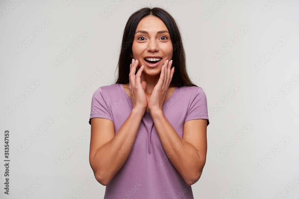 Surprised young attractive brunette lady with casual hairstyle looking at camera with wide eyes and mouth opened, keeping hands on her face while standing over white background Surprised young attractive brunette lady with casual hairstyle looking at camera with wide eyes and mouth opened, keeping hands on her face while standing over white background