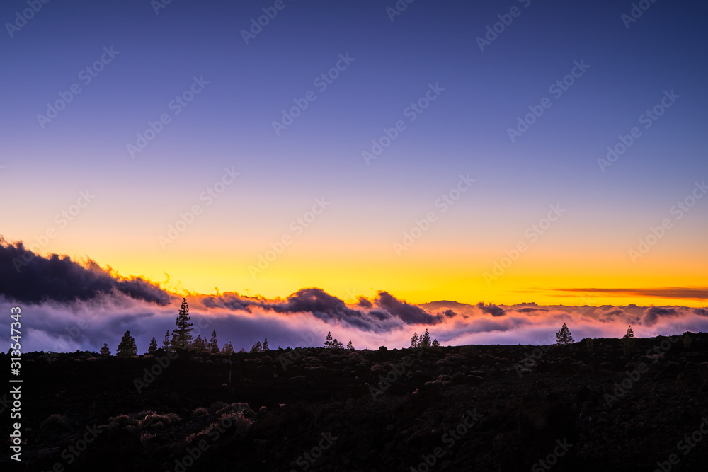 Spain, Tenerife, Orange sky and moonlight shining on arid black lava nature landscape above the clouds in mountains of caldera after sunset by night