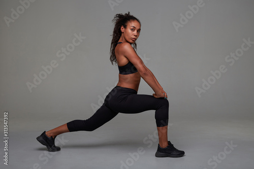 Fitness young attractive dark skinned curly model stretching her legs and looking at camera with folded lips, wanting to be fit and healthy, posing over grey background