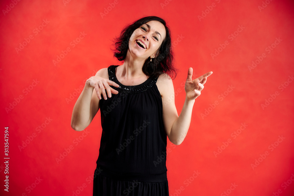 Portrait of a pretty brunette woman in a black dress on a red background. Shows vivid emotions with hands in different poses right in front of the camera.