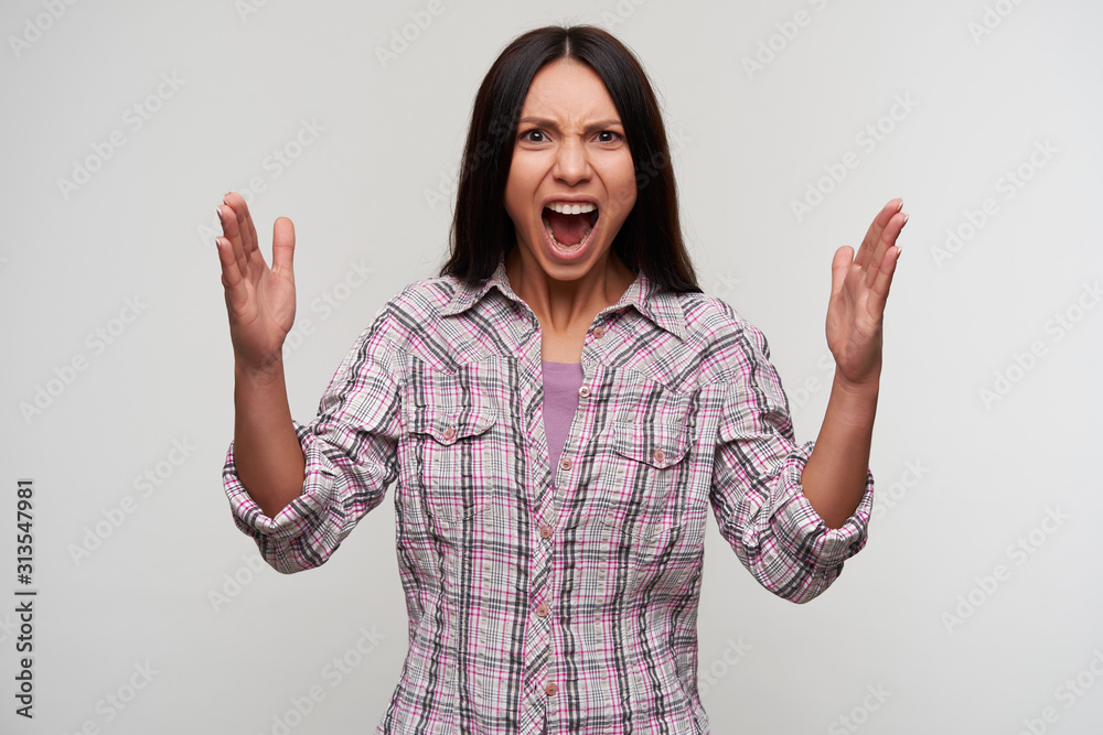 Excited young pretty dark haired female with casual hairstyle looking emotionally to camera with raised hands and keeping mouth wide opened, standing against white background Excited young pretty dark haired female with casual hairstyle looking emotionally to camera with raised hands and keeping mouth wide opened, standing against white background