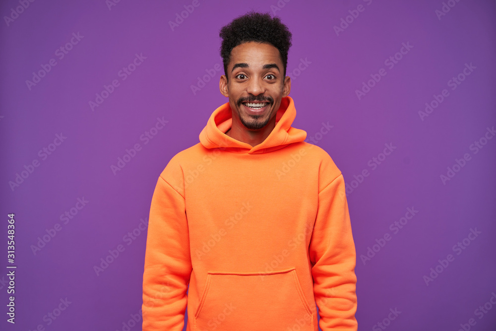 Joyful young lovely brown-eyed dark skinned brunette male with beard looking surprisedly at camera and smiling widely while standing over purple background in sporty wear Joyful young lovely brown-eyed dark skinned brunette male with beard looking surprisedly at camera and smiling widely while standing over purple background in sporty wear