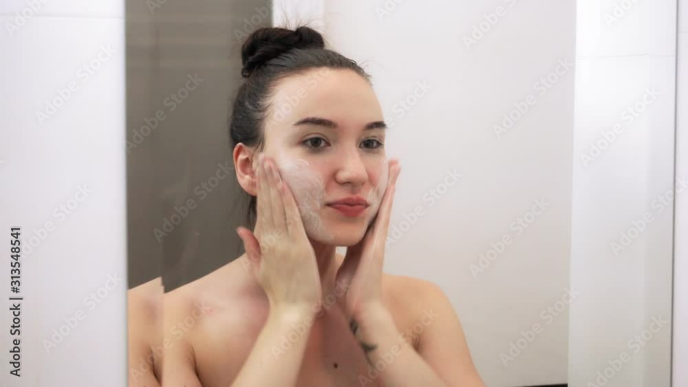 Happy attractive young woman washing her face with cleansing foam. Close-up view reflection in the mirror. Happy attractive young woman washing her face with cleansing foam. Close-up view reflection in the mirror.