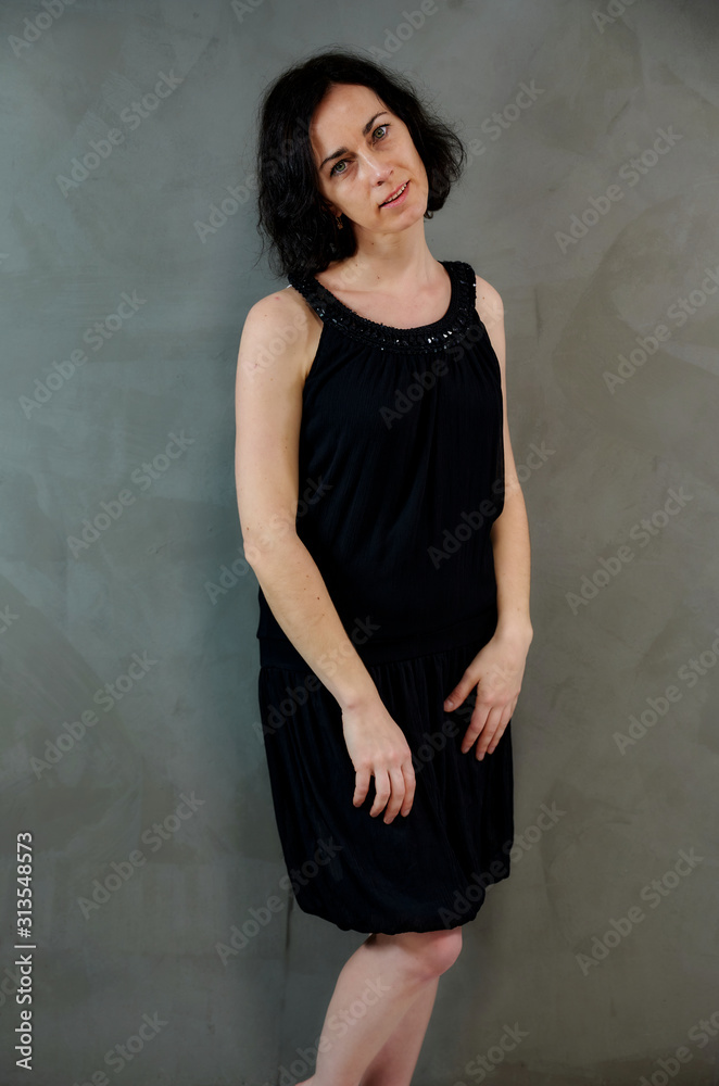 Full length portrait of a pretty emotional brunette woman in black dress on alternative gray background in vintage interior. Standing right in front of the camera.