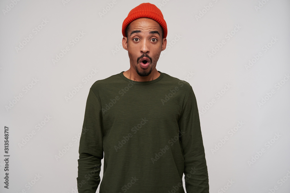 Overwhelmed young attractive bearded brunette male with dark skin looking amazedly at camera with round eyes, wearing red hat and khaki sweater over white background Overwhelmed young attractive bearded brunette male with dark skin looking amazedly at camera with round eyes, wearing red hat and khaki sweater over white background