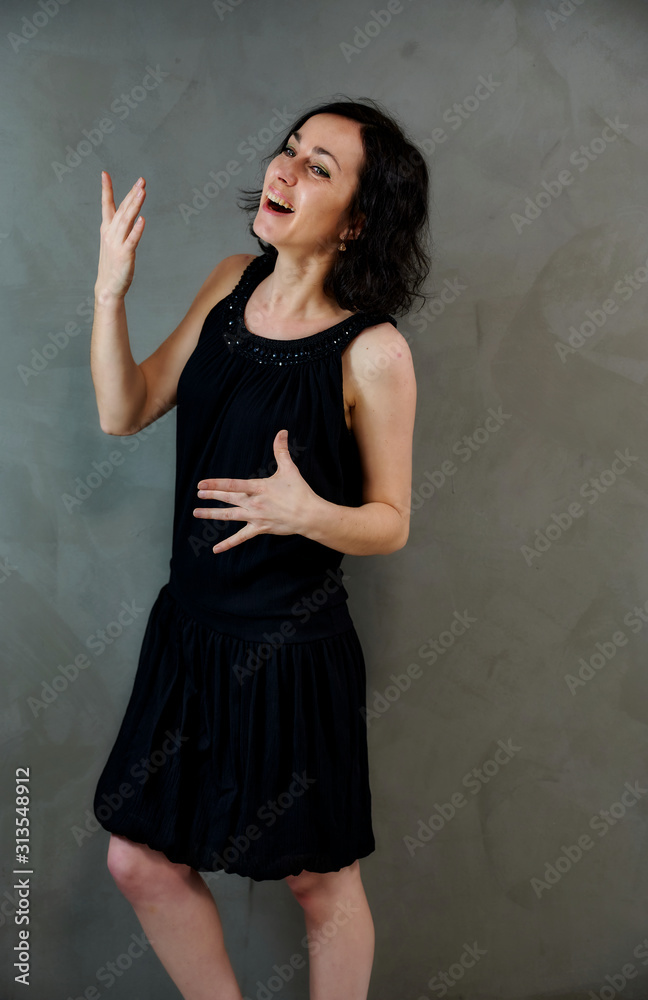 Full length portrait of a pretty emotional brunette woman in black dress on alternative gray background in vintage interior. Standing right in front of the camera. Full length portrait of a pretty emotional brunette woman in black dress on alternative gray background in vintage interior. Standing right in front of the camera.