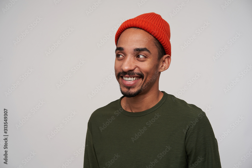 Close-up of charming young positive dark haired bearded man with dark skin looking happily aside with wide sincere smile, being in good mood while standing against white background