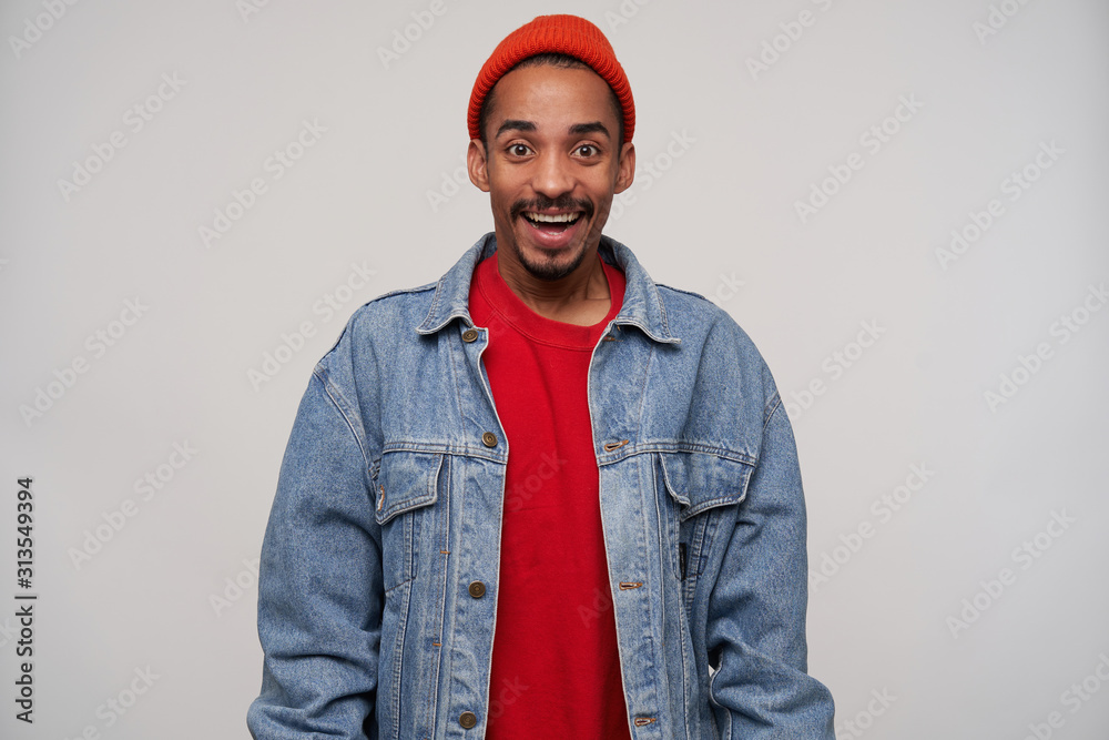 Indoor shot of cheerful attractive young brown-eyed bearded dark skinned guy looking happily to camera with wide smile, being in high spirit while standing over white background Indoor shot of cheerful attractive young brown-eyed bearded dark skinned guy looking happily to camera with wide smile, being in high spirit while standing over white background
