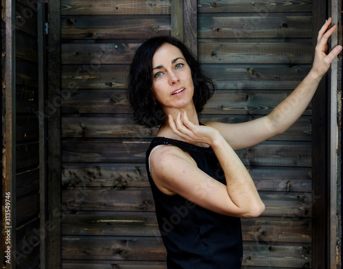 Portrait of a pretty smiling brunette woman in black dress on wooden background in vintage interior. Standing right in front of the camera with vivid emotions. © Вячеслав Чичаев