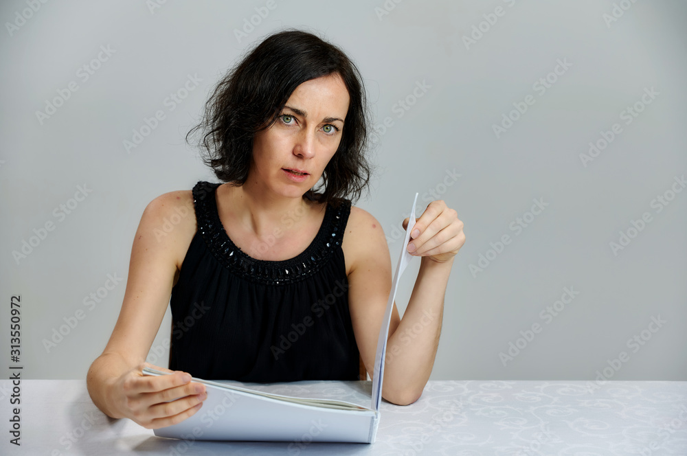 Portrait of a cute smiling talking brunette woman in a black dress on a white background. Sits at a table right in front of the camera with vivid emotions with a folder in his hands. Portrait of a cute smiling talking brunette woman in a black dress on a white background. Sits at a table right in front of the camera with vivid emotions with a folder in his hands.