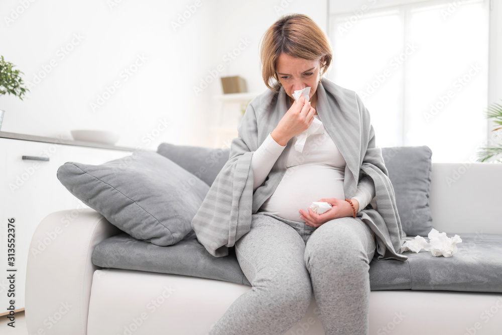 Pregnant woman catching cold, flu, virus, sitting at home on a couch, blowing nose and being sick. Pregnant woman catching cold, flu, virus, sitting at home on a couch, blowing nose and being sick.