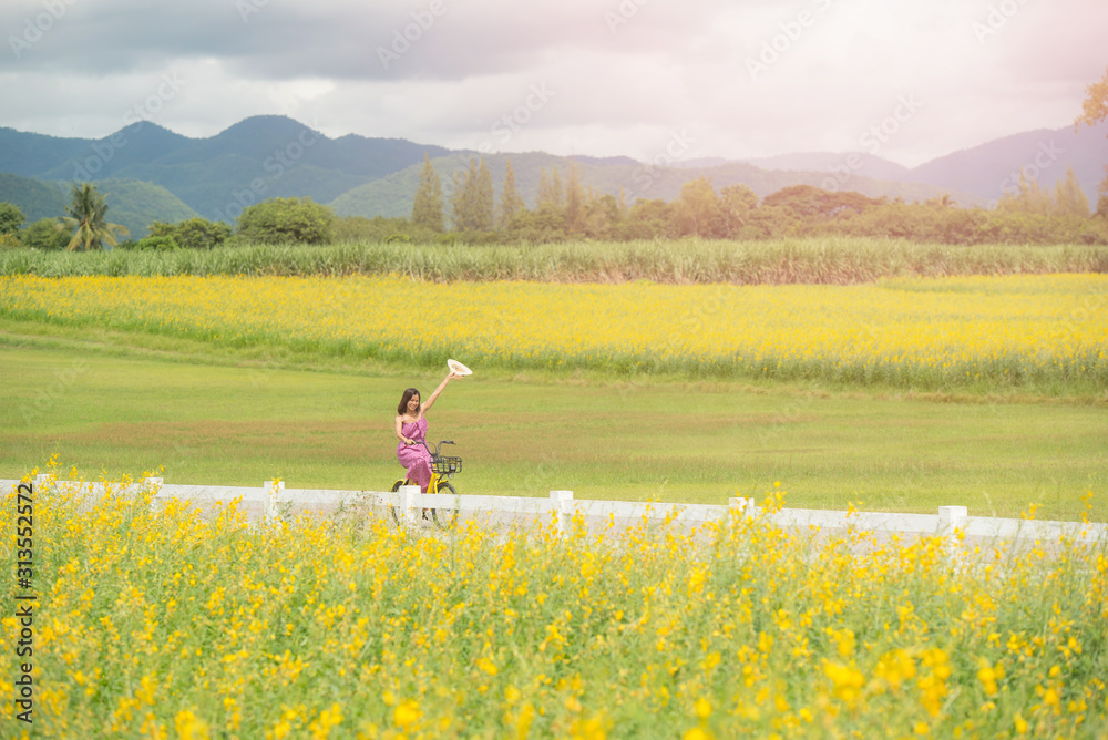 asia woman with a hat in her hand walks in a field with field flowers and smiles sincerely, happy enjoying summer in yellow field at sunset. woman riding bicycle in flower field. concept of freedom.