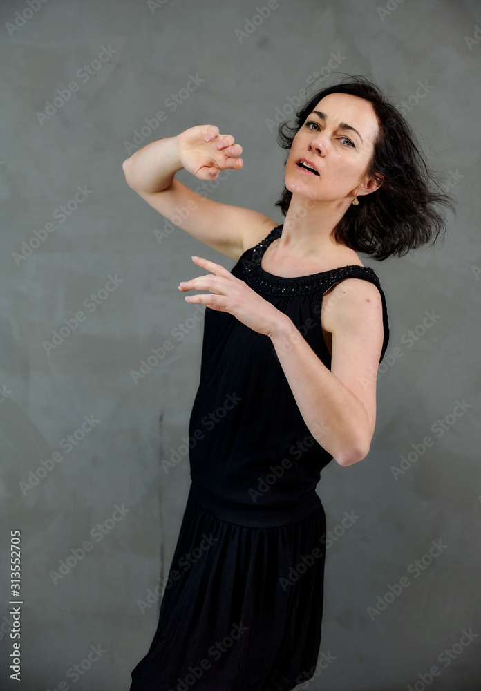 Model Standing right in front of the camera with vivid emotions. Vertical portrait of a cute smiling brunette woman in black dress on gray alternative background.