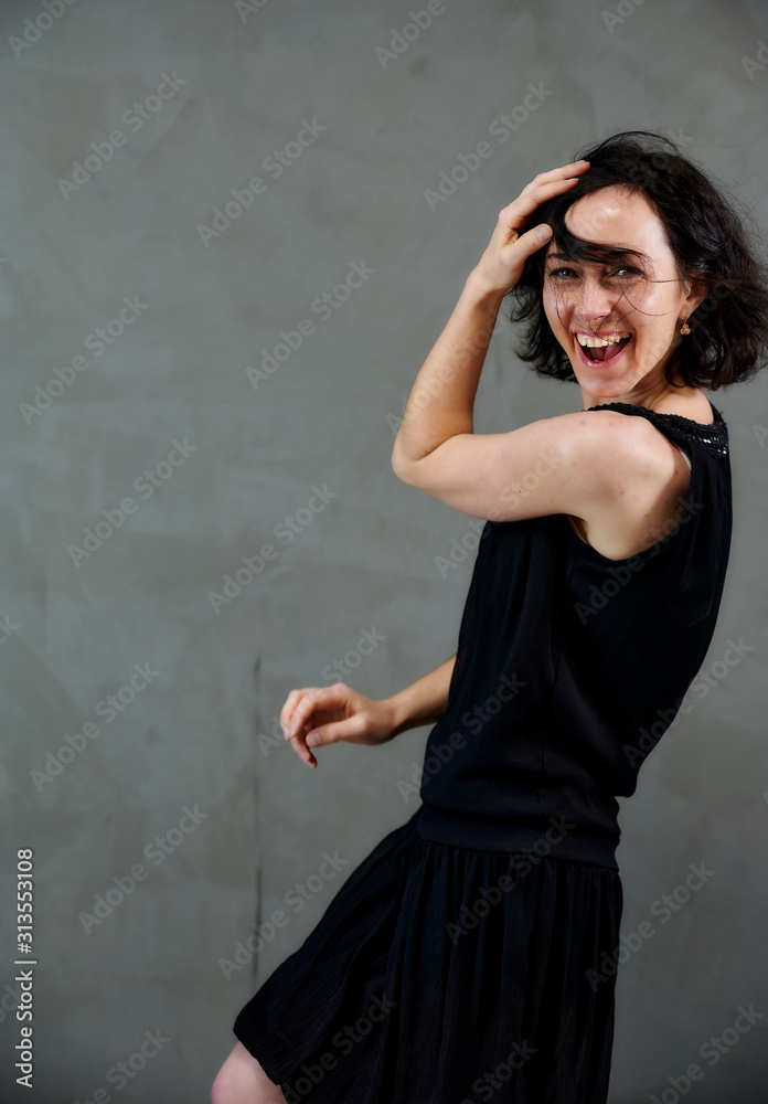 Model Standing right in front of the camera with vivid emotions. Vertical portrait of a cute smiling brunette woman in black dress on gray alternative background. Model Standing right in front of the camera with vivid emotions. Vertical portrait of a cute smiling brunette woman in black dress on gray alternative background.