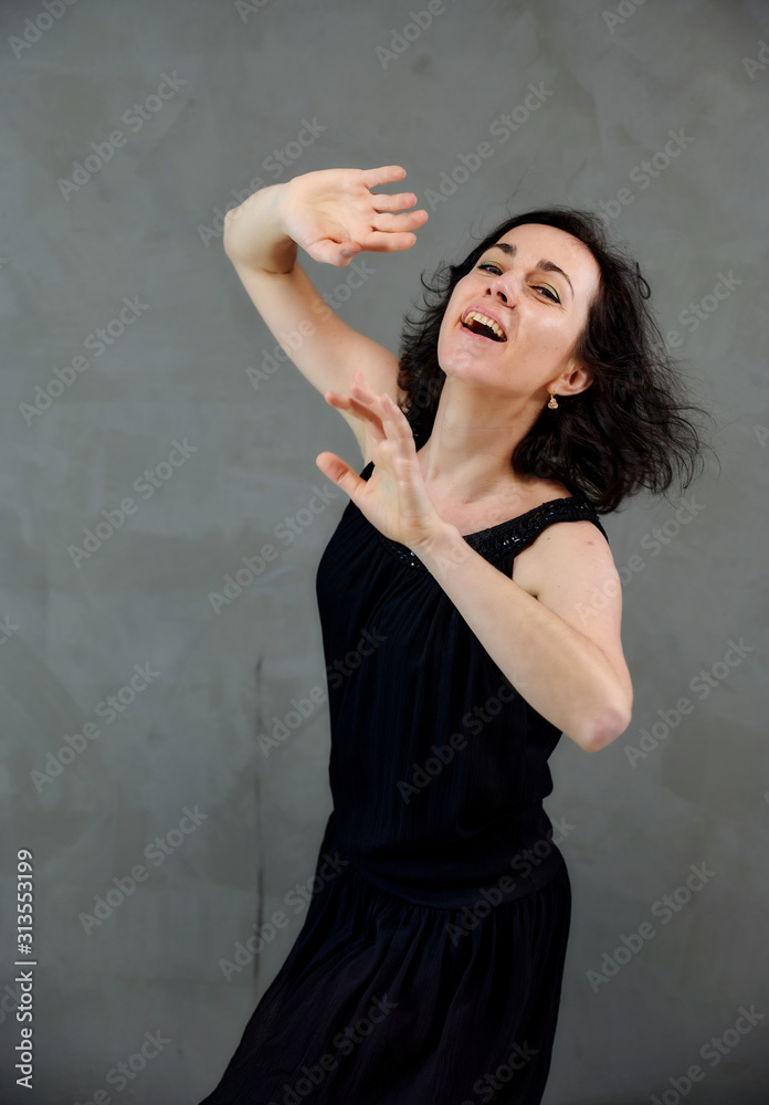 Model Standing right in front of the camera with vivid emotions. Vertical portrait of a cute smiling brunette woman in black dress on gray alternative background. Model Standing right in front of the camera with vivid emotions. Vertical portrait of a cute smiling brunette woman in black dress on gray alternative background.