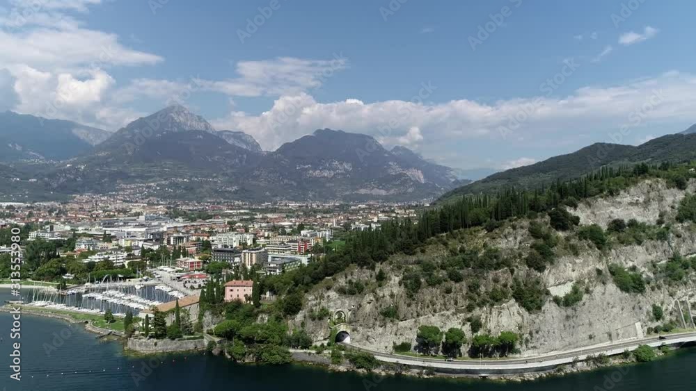 Aerial view of Riva del Garda lake shore mountain showing road and tunnel located next to bright blue fresh water this town is a popular tourist attraction in summer holidays 4k high resolution Aerial view of Riva del Garda lake shore mountain showing road and tunnel located next to bright blue fresh water this town is a popular tourist attraction in summer holidays 4k high resolution