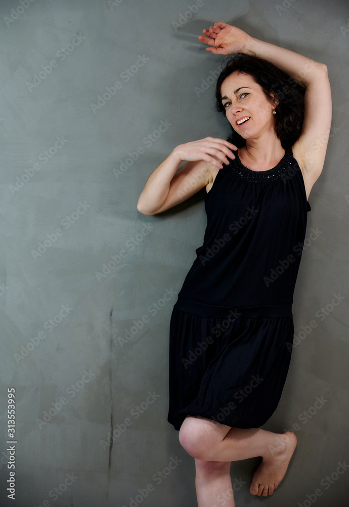 Model Standing right in front of the camera with vivid emotions. Vertical portrait of a cute smiling brunette woman in black dress on gray alternative background. Model Standing right in front of the camera with vivid emotions. Vertical portrait of a cute smiling brunette woman in black dress on gray alternative background.