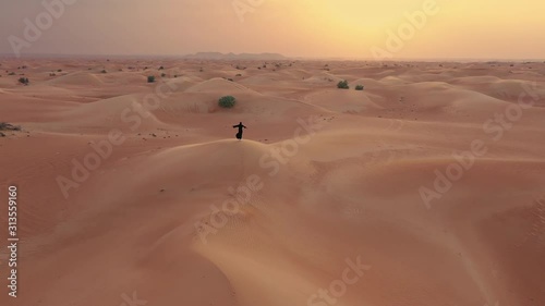 AERIAL. Camera moving around woman in traditional Emirati dress walking in a desert in strog wind and sunset. photo